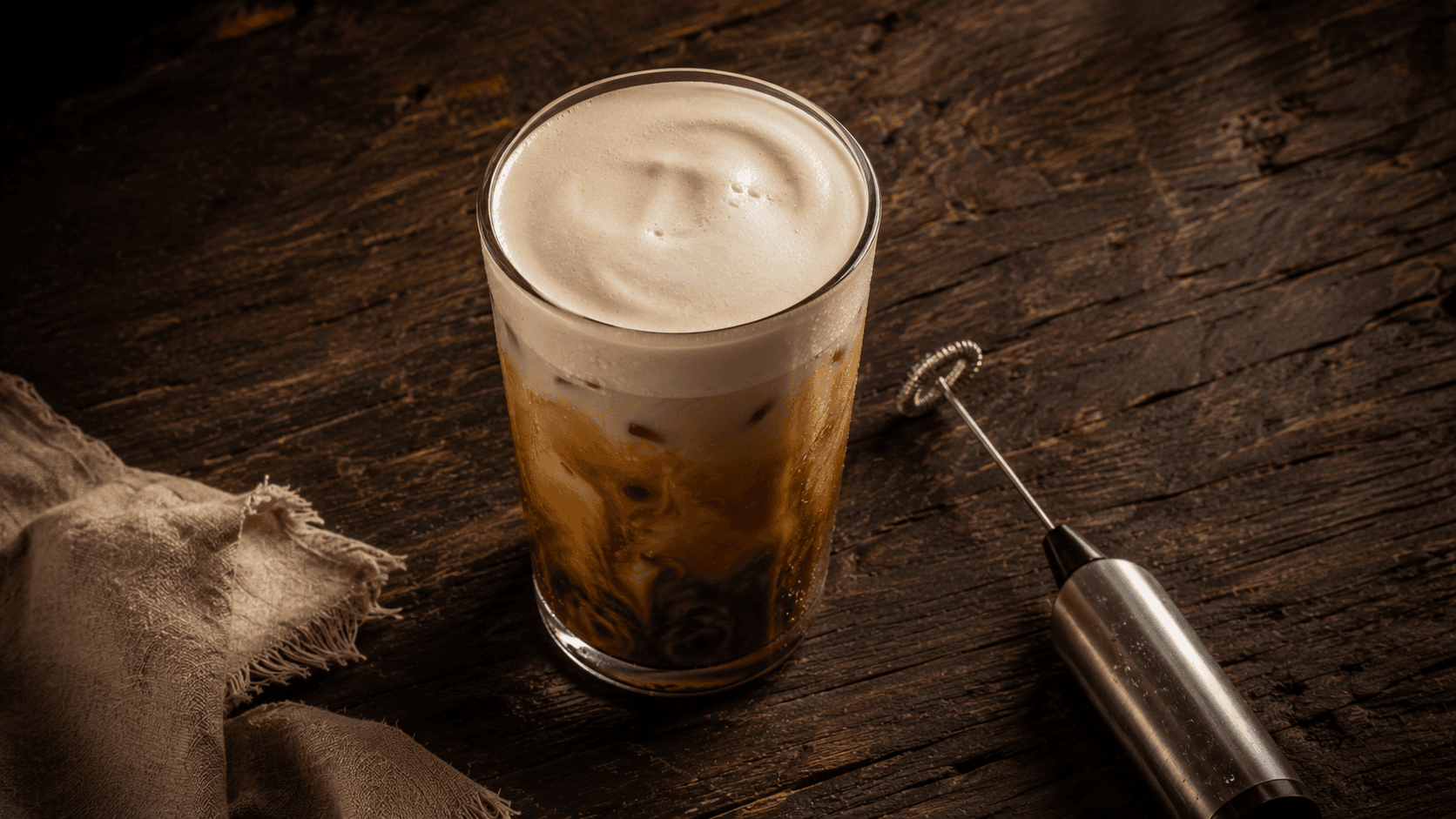 Overhead shot of iced coffee topped with thick cold foam next to a handheld milk frother on a dark wooden table