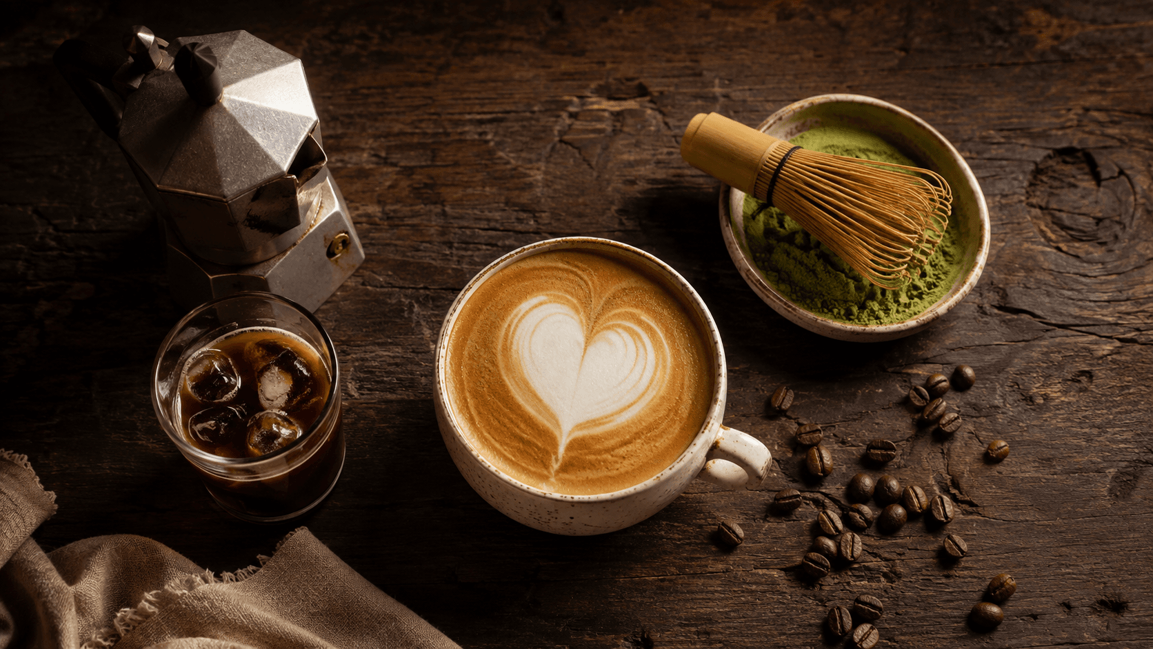 Overhead shot of a latte with a heart, a moka pot, iced coffee, matcha with a chasen whisk, and scattered coffee beans on a dark wooden table